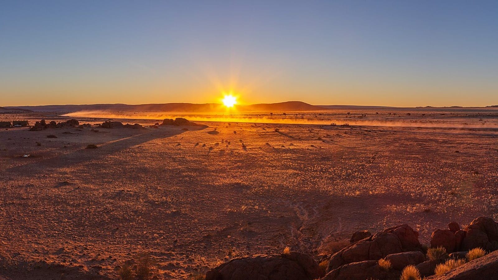 Low sun over a stony Namib plain with sparse grass tufts and red rock outcrops, Namibia.