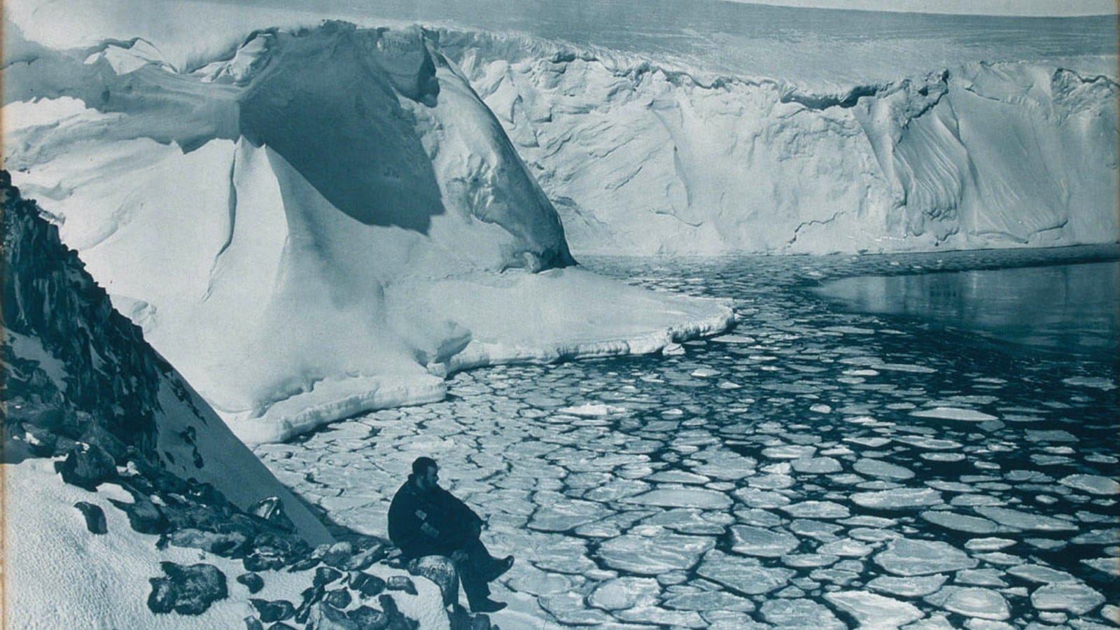 Glacier ice cliff above pancake ice with a seated man on the rocky shore, Commonwealth Bay, Antarctica.