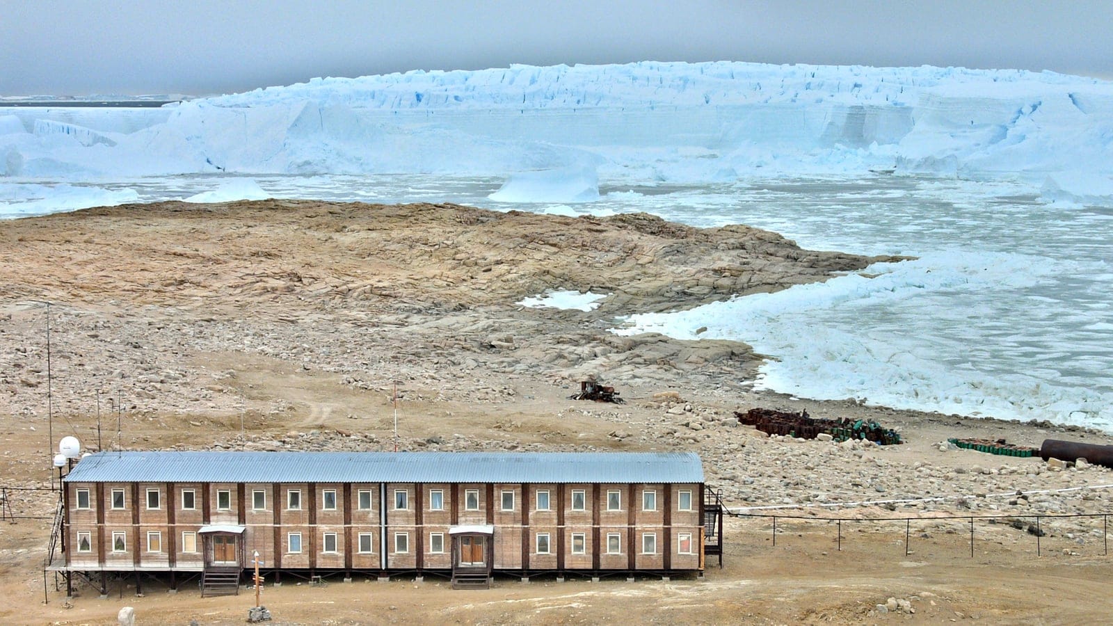 A stilted station building on bare rock above sea ice and an ice cliff at Progress Station, East Antarctica.