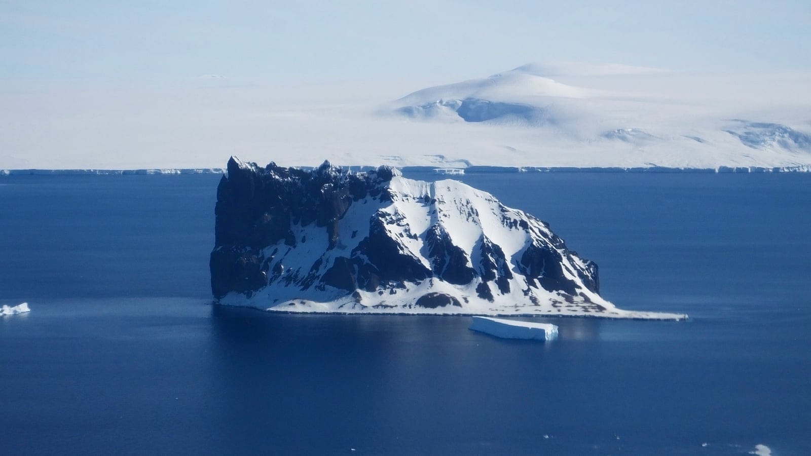 Aerial view of Sims Island, a snow- and ice-covered Antarctic island rising from sea ice and dark open water off the Palmer Land coast.