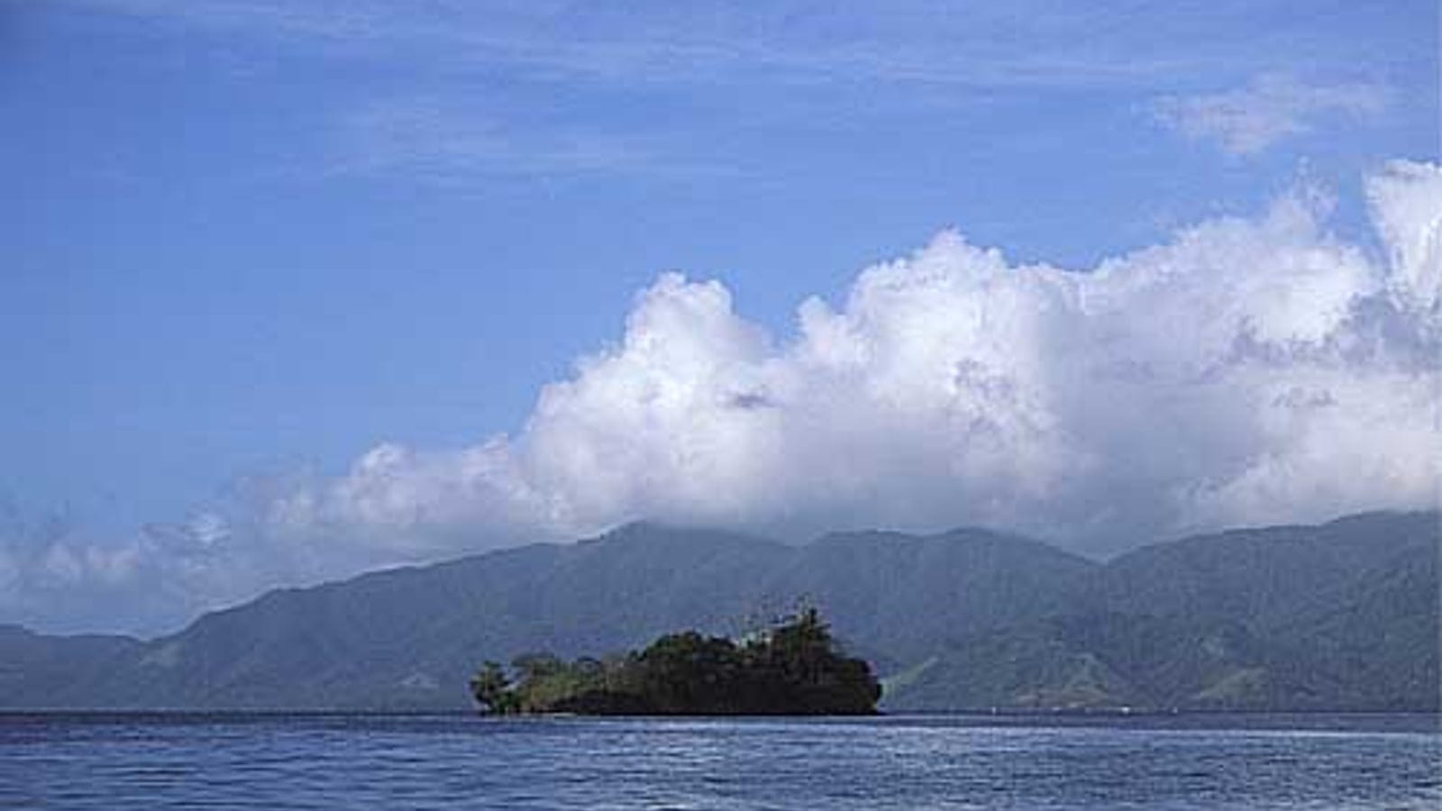 Small forested islet and hazy ridges of Fergusson Island under towering cumulus, D'Entrecasteaux Islands.