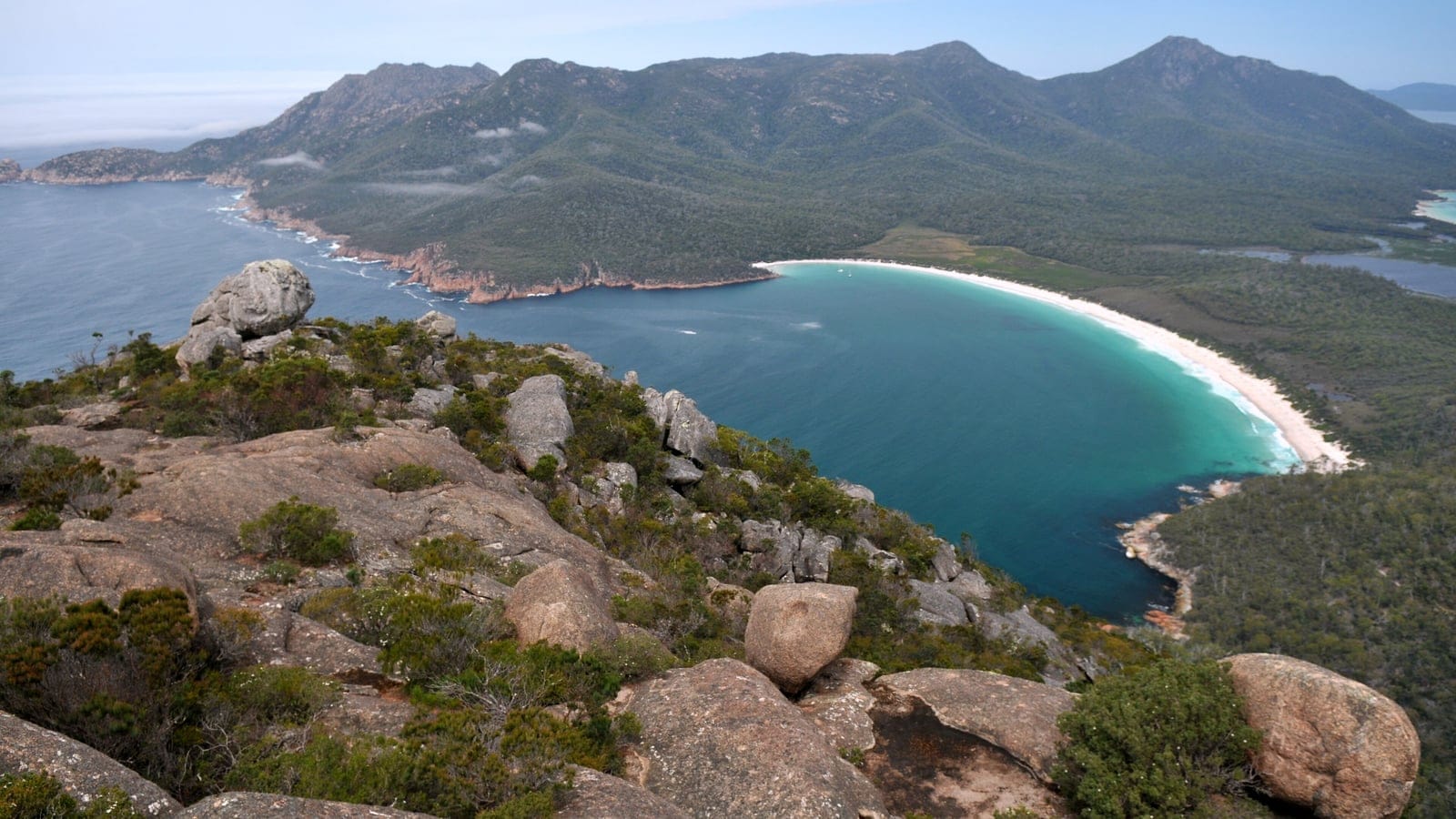 Granite boulders and low coastal heath above Wineglass Bay, with forested hills behind, Freycinet, Tasmania.