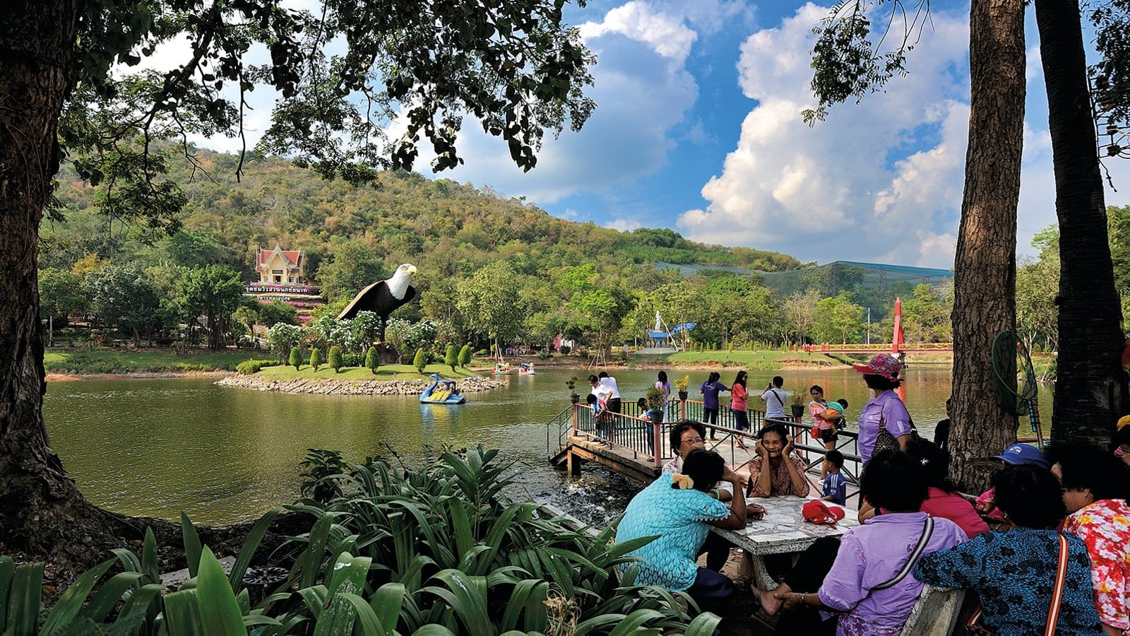 Visitors at tables beside an ornamental lake below wooded hills at Chai Nat Bird Park, Thailand.