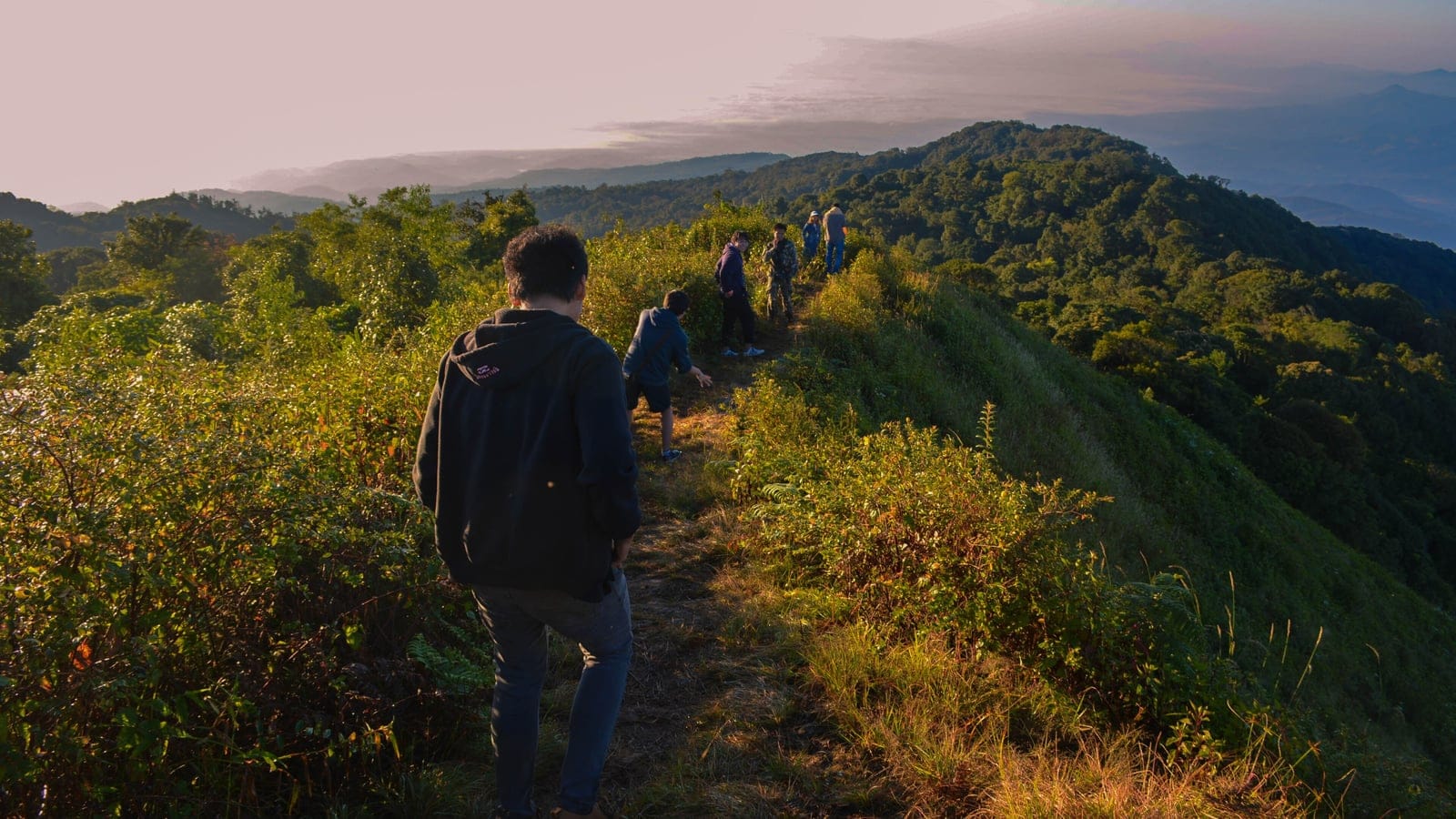 Hikers on a grassy ridge trail above forested hills at Doi Pha Hom Pok, Thailand.