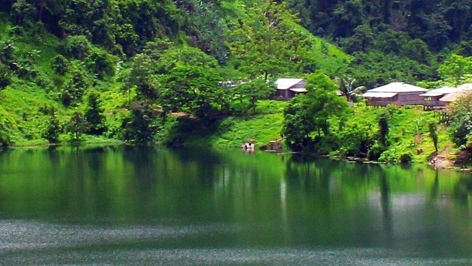 Steep green slopes and stilt houses reflected in the still water of Boga Lake, Chittagong Hill Tracts, Bangladesh.