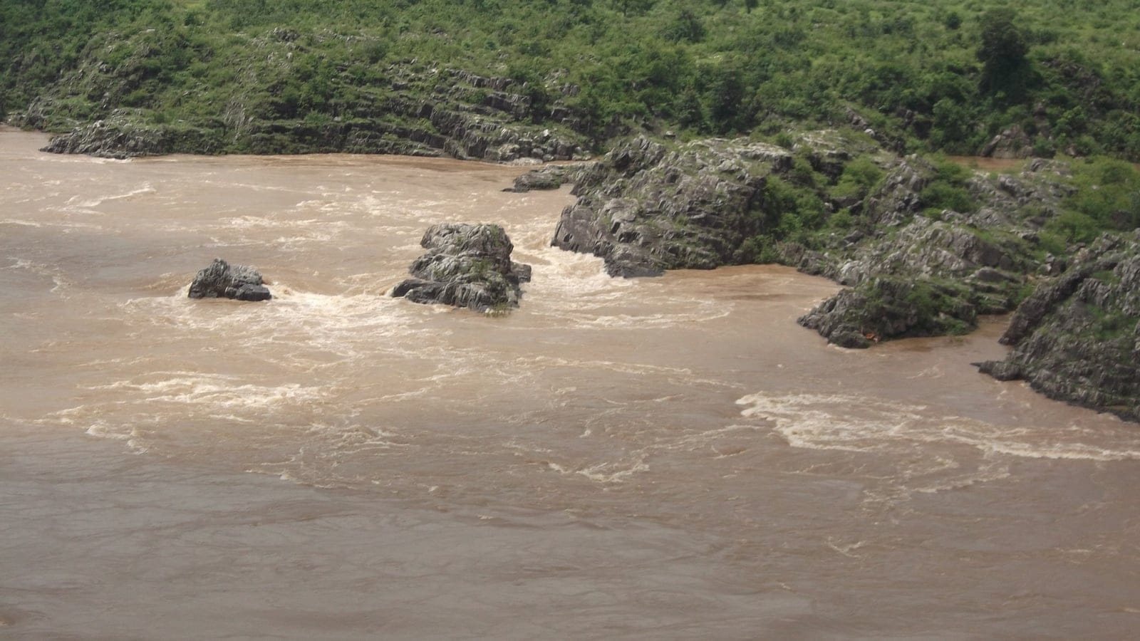 El río Narmada, crecido por el monzón, rompiendo sobre repisas de roca bajo orillas cubiertas de matorral, Bhedaghat, India.