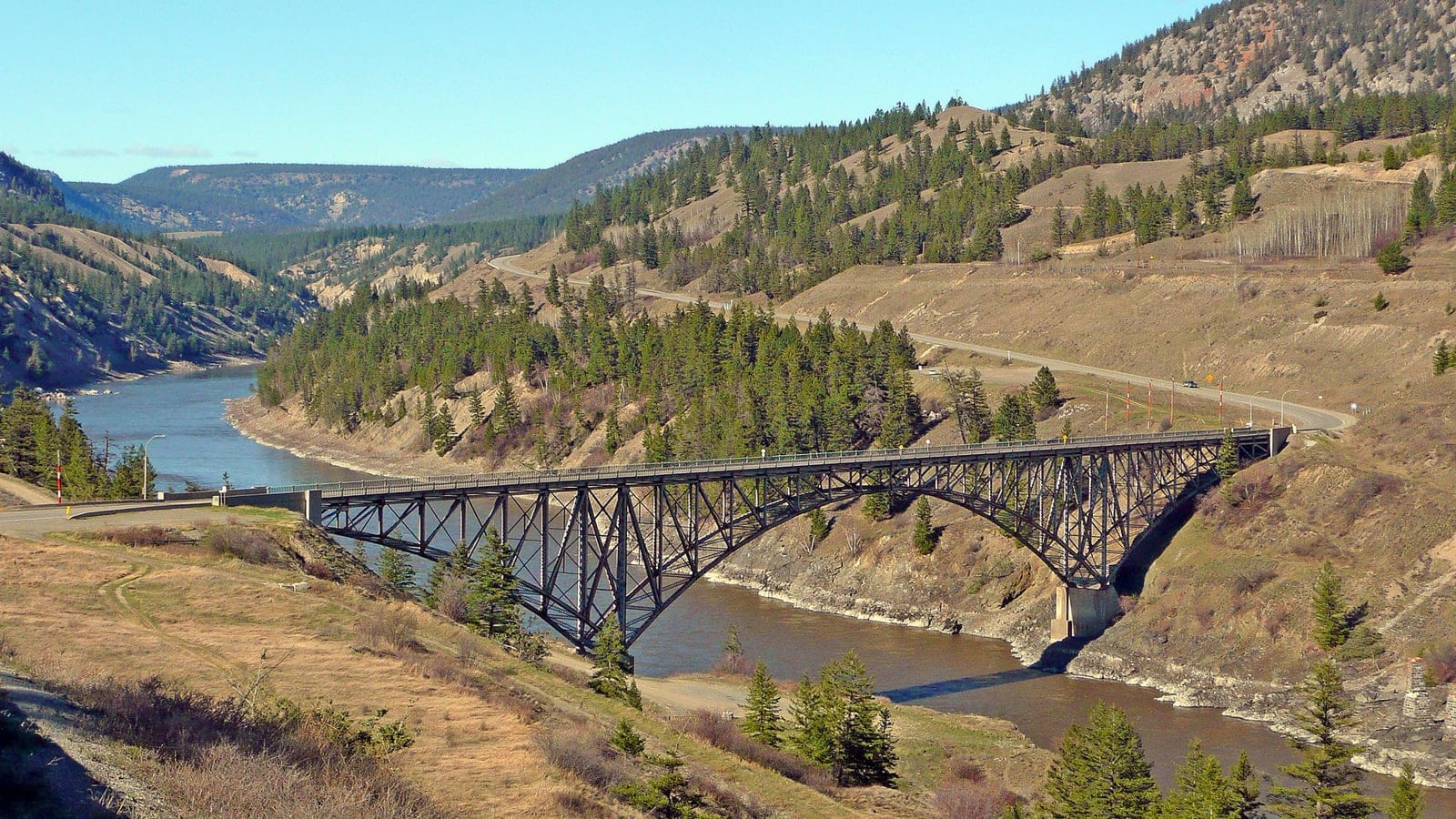 A steel truss bridge crossing the Fraser River between conifer dotted canyon slopes, British Columbia.