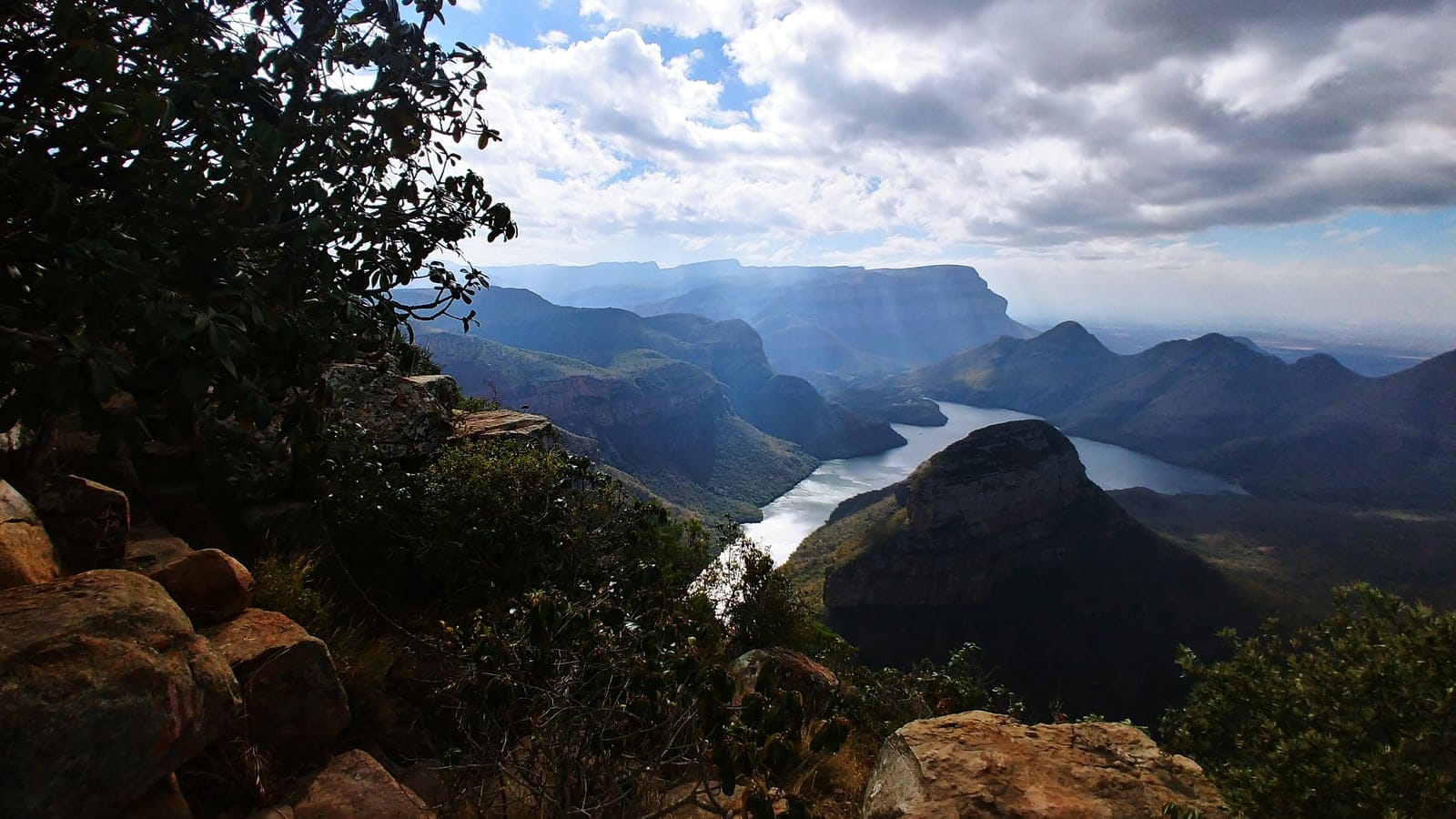 Sweeping view of the Blyde River Canyon on the eastern Great Escarpment, with steep cliff-faced mountains dropping toward a river valley and subtropical escarpment vegetation under a cloudy sky.