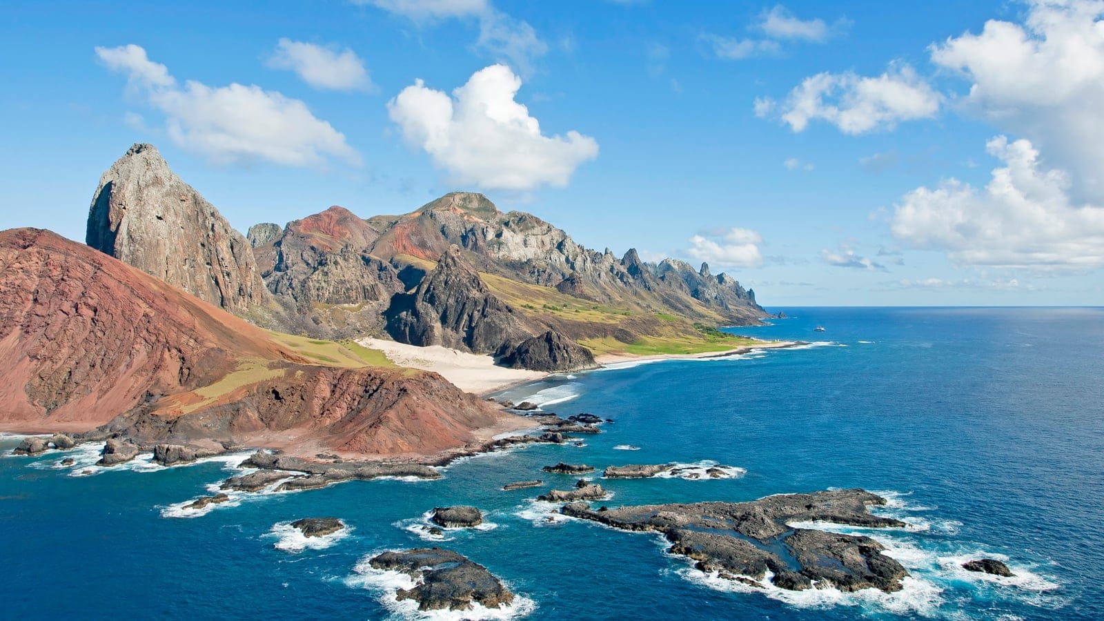 Eroded red volcanic slopes with thin grass cover above surf-washed reefs at Trindade Island, Brazil.