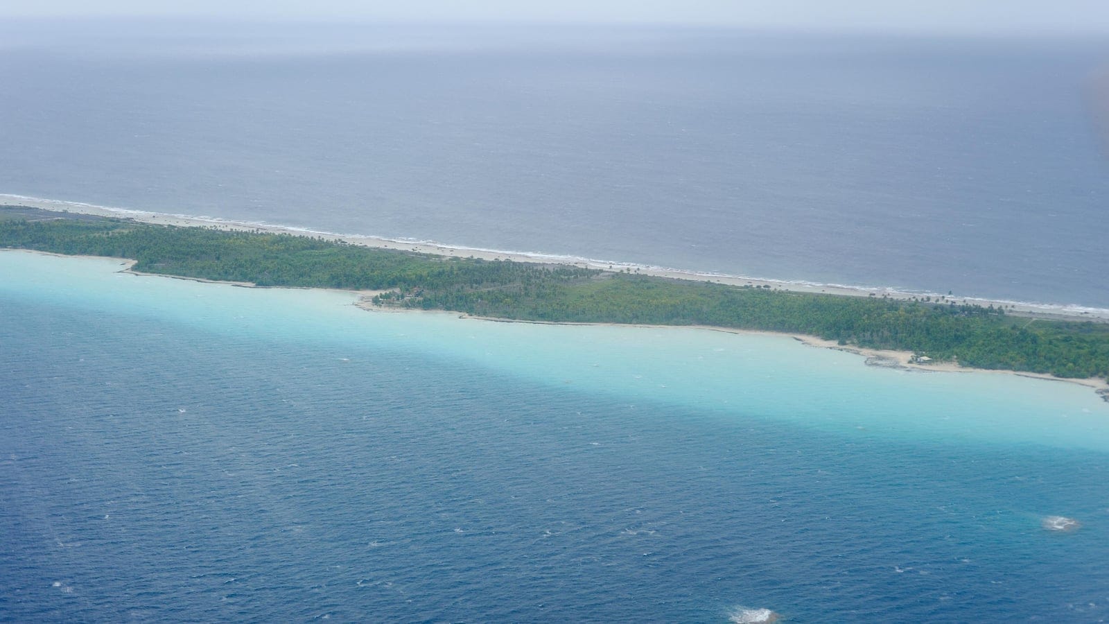Aerial view of a narrow forested motu between a turquoise reef lagoon and open ocean, Tuamotu Archipelago.