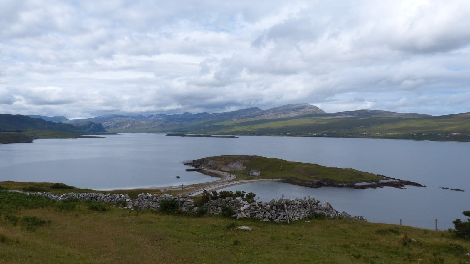 Sheep-grazed grassland and a tumbled drystone wall above a sea loch and its tied island at Heilam, Scotland.