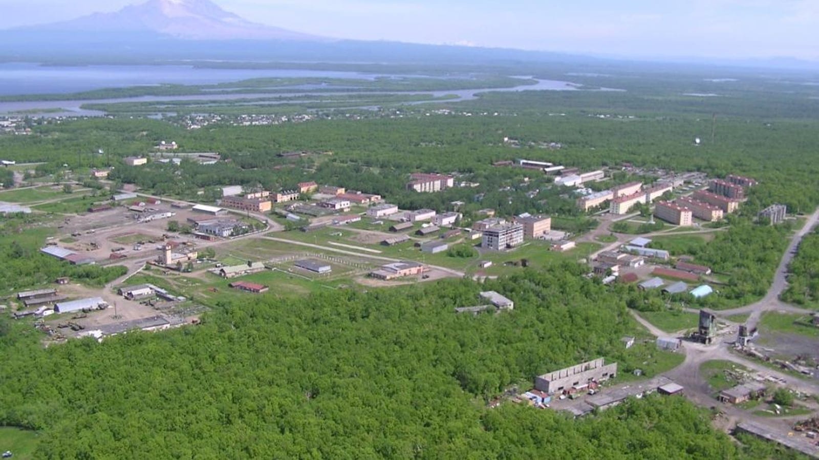 Bloques de apartamentos y patios industriales rodeados por un denso bosque de abedules bajo un volcán nevado en Klyuchi, Kamchatka.