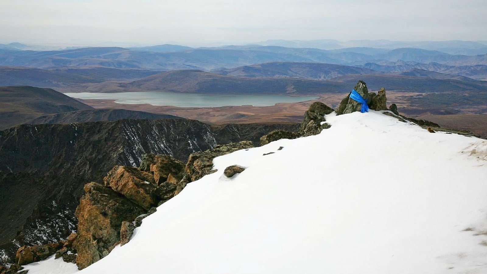 Snow-covered summit ridge and a cairn with a blue khadag above a pale lake, Khangai Mountains, Mongolia.