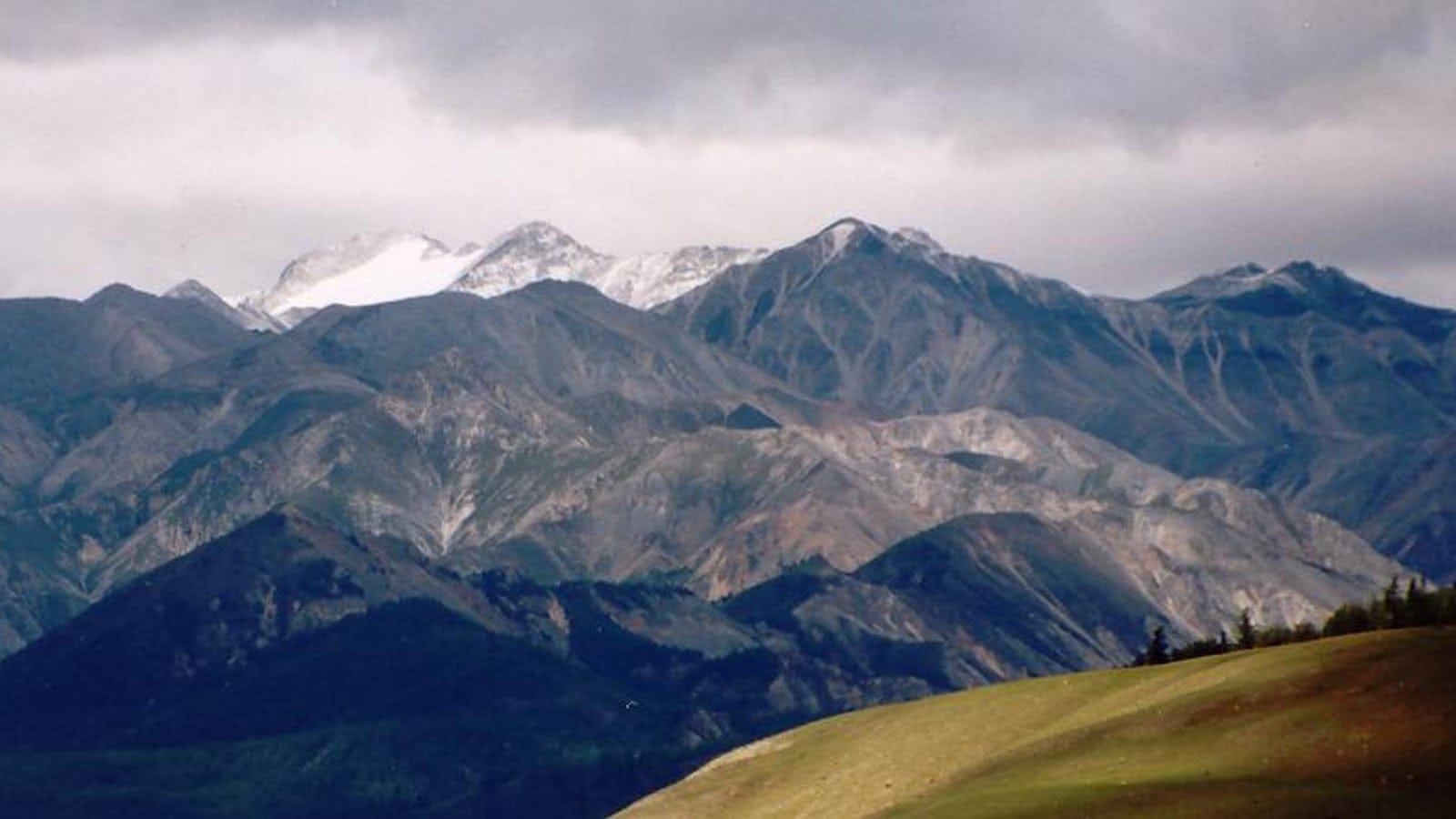 Grey scree ridges and a green alpine meadow slope under storm cloud, Eastern Sayan, Russia.