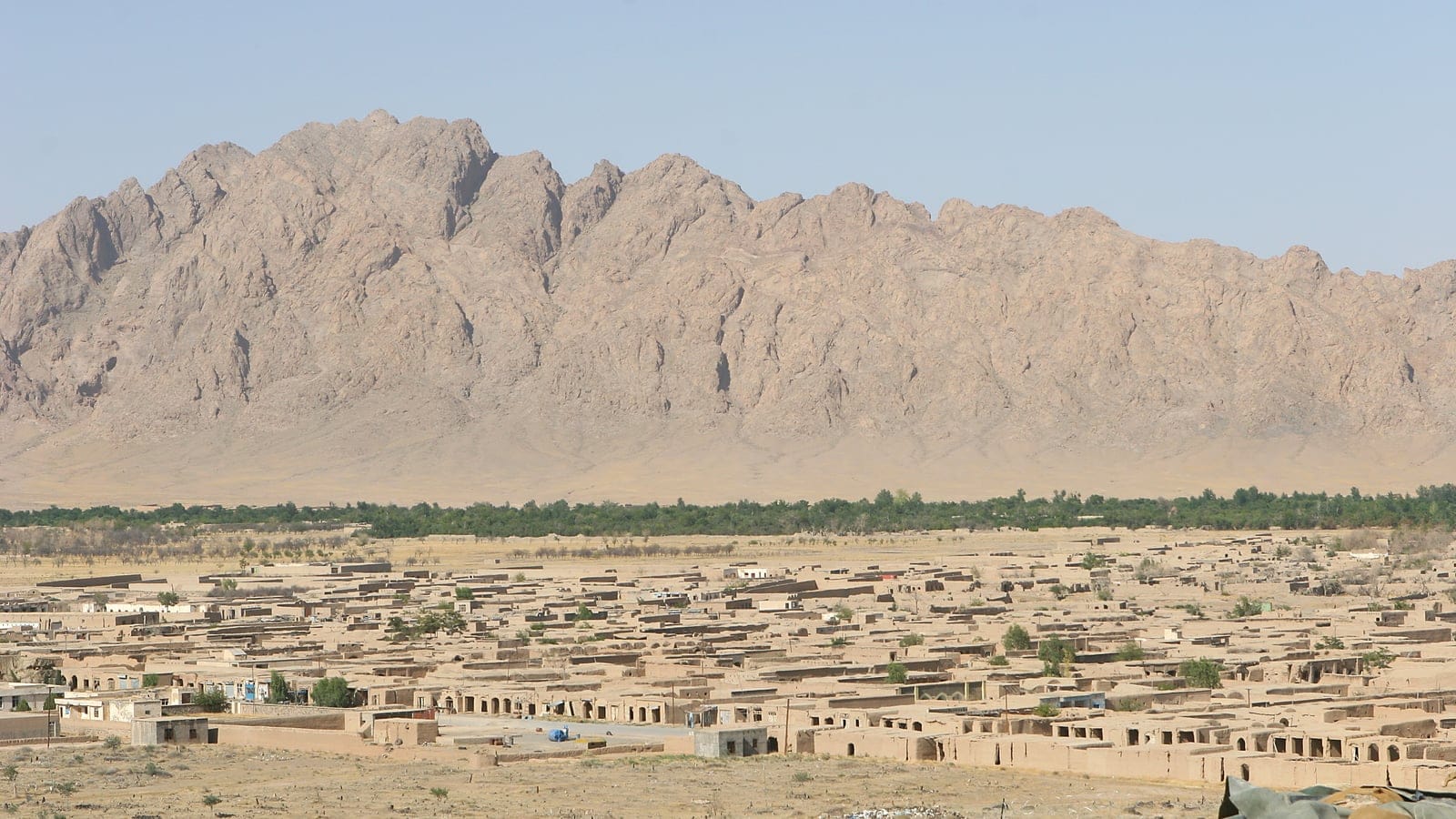 Flat-roofed mud-brick compounds and a green tree belt below a bare rock ridge at Now Zad, Helmand, Afghanistan.