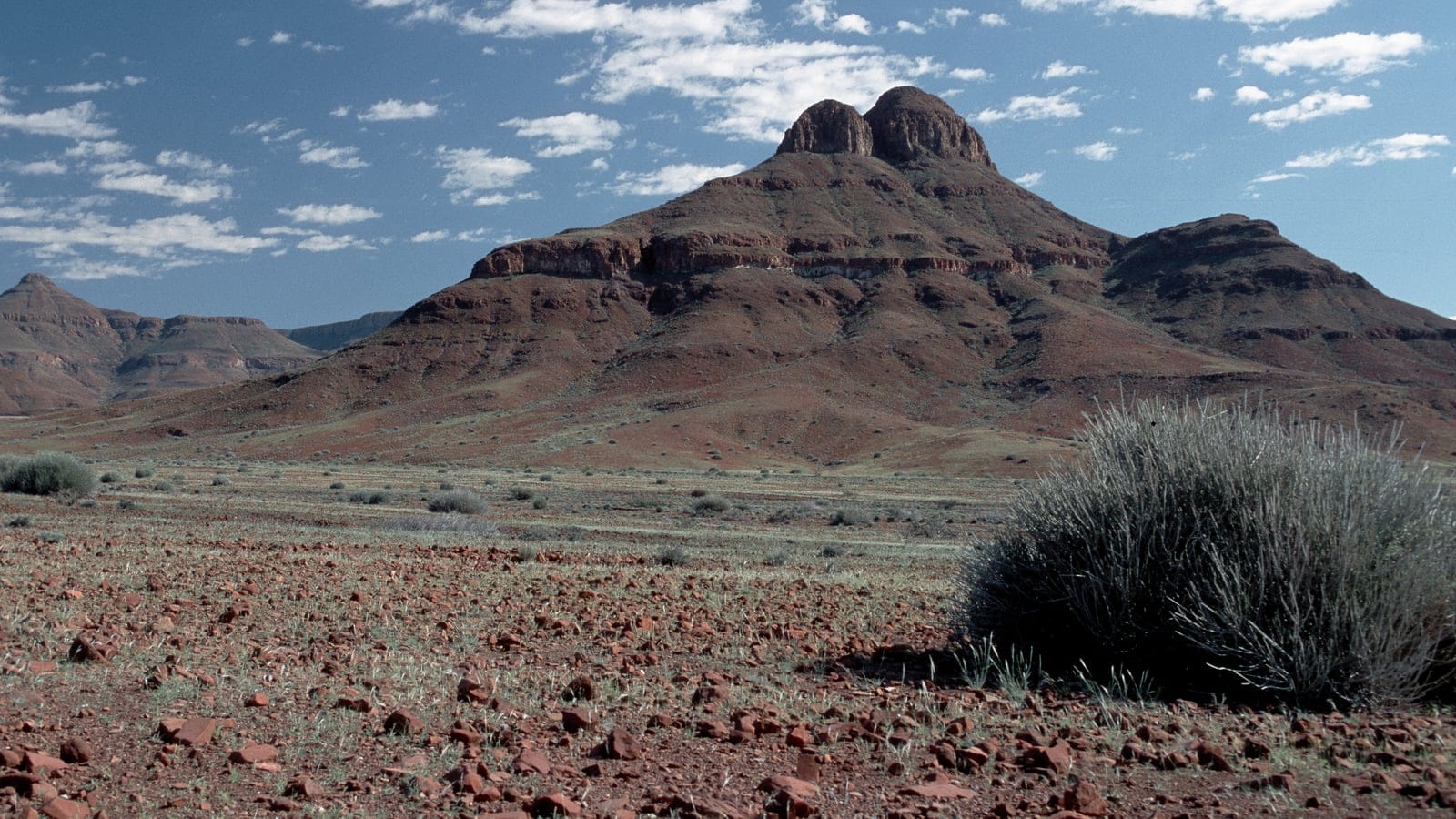 Sparse grass and grey shrubs on a red stony plain below a banded sandstone massif north of Orupembe, Namibia.
